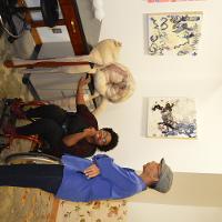 A man in sandals and a woman in a wheelchair next to a wall of paintings 