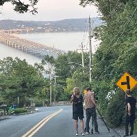 Two people standing on a road overlooking a bridge and river at dusk, with a yellow warning sign nearby.