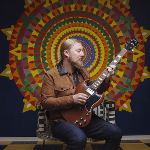 Guitarist kneeling while playing an electric guitar in front of a large colorful mandala-style mural.