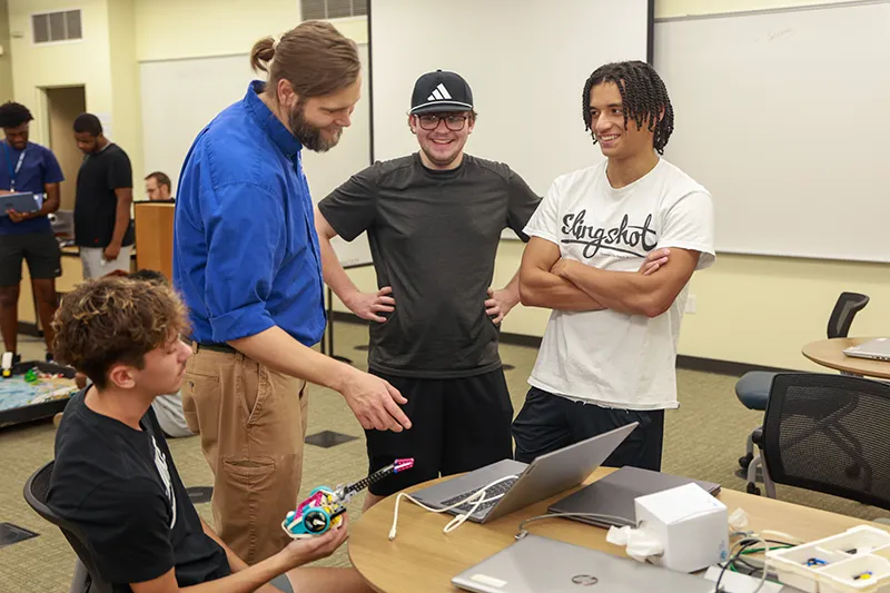 An instructor and three students laughing together around a laptop and electronics project in a lab