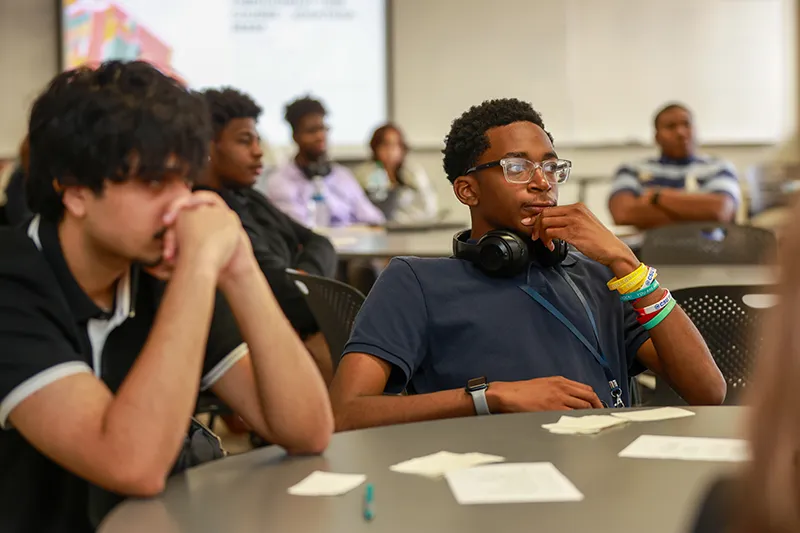 Students sitting at tables in a classroom, listening attentively to a presentation