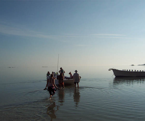 Two small wooden boats, one with several people wading towards it, one empty. Dim blue sky.