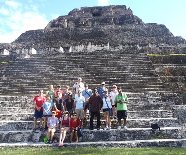 Lots of students smiling and posing at the base of a huge rock monument, which dwarfs them