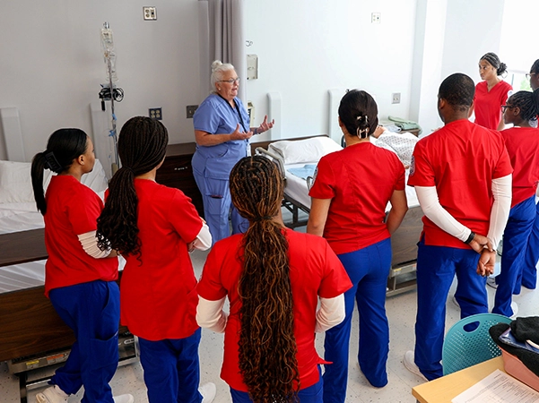 A group of nursing students surrounding a bed and paying attention to a nurse as she teaches them.