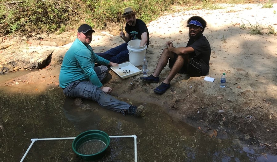Three male students sitting on the edge of a creek gathering rocks