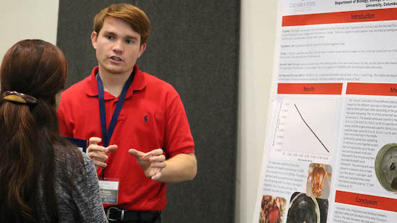 A male student standing in front of a poster talking to another individual