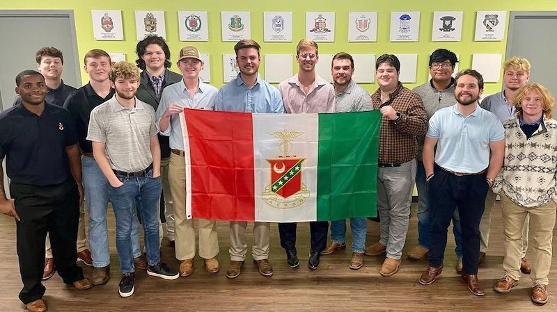 Fraternity members posing inside, two of whom holding a red, white, and green flag