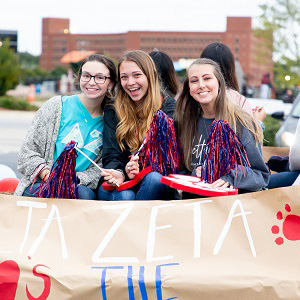 Three students smiling on a parade float holding pom-poms and a banner